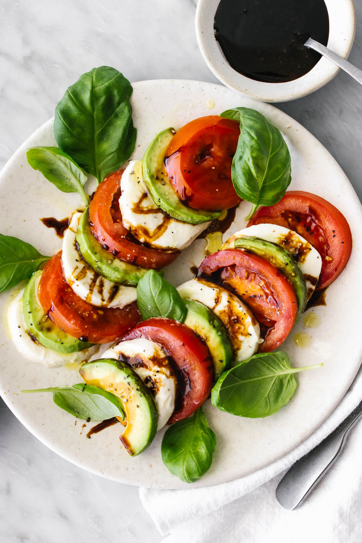 Avocado caprese salad on a white plate next to balsamic vinegar.