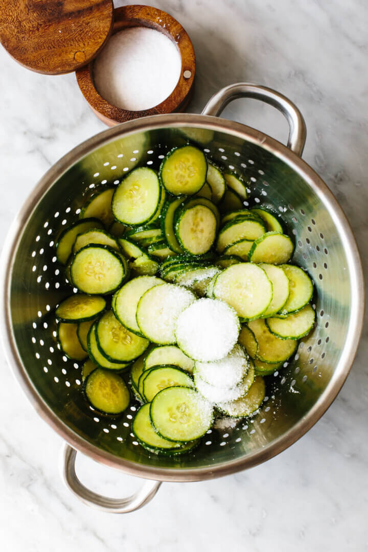 Adding salt to cucumbers in a colander for cucumber salad