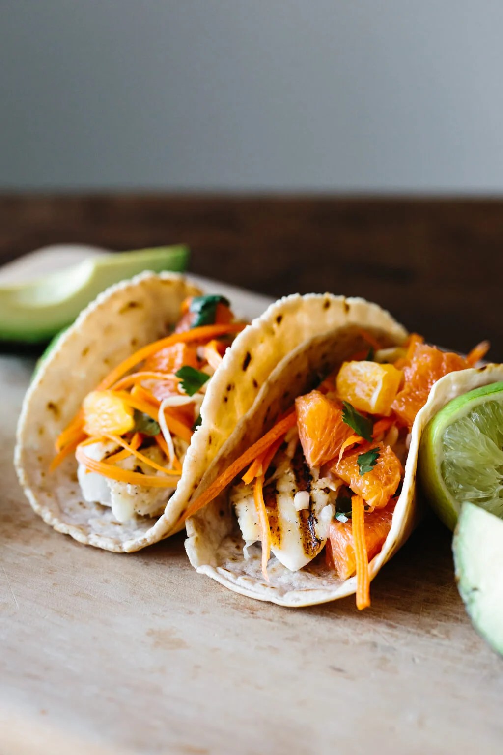 Two fish tacos on a table next to limes and avocado.