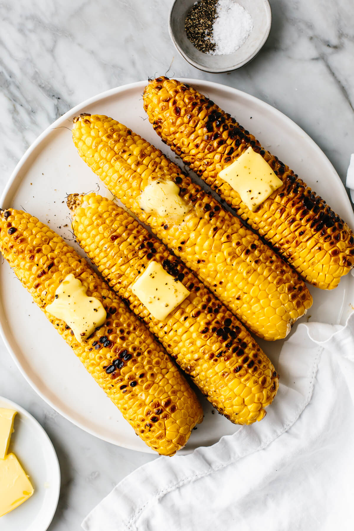 Grilled corn on the cobs on a white plate next to butter.