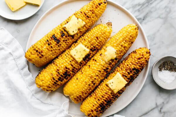 A circle plate with grilled corn on the cobs next to salt and pepper.