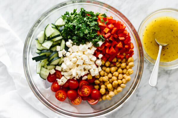 Ingredients for a Mediterranean chickpea salad in a glass bowl next to a small bowl of lemon vinaigrette on a table.