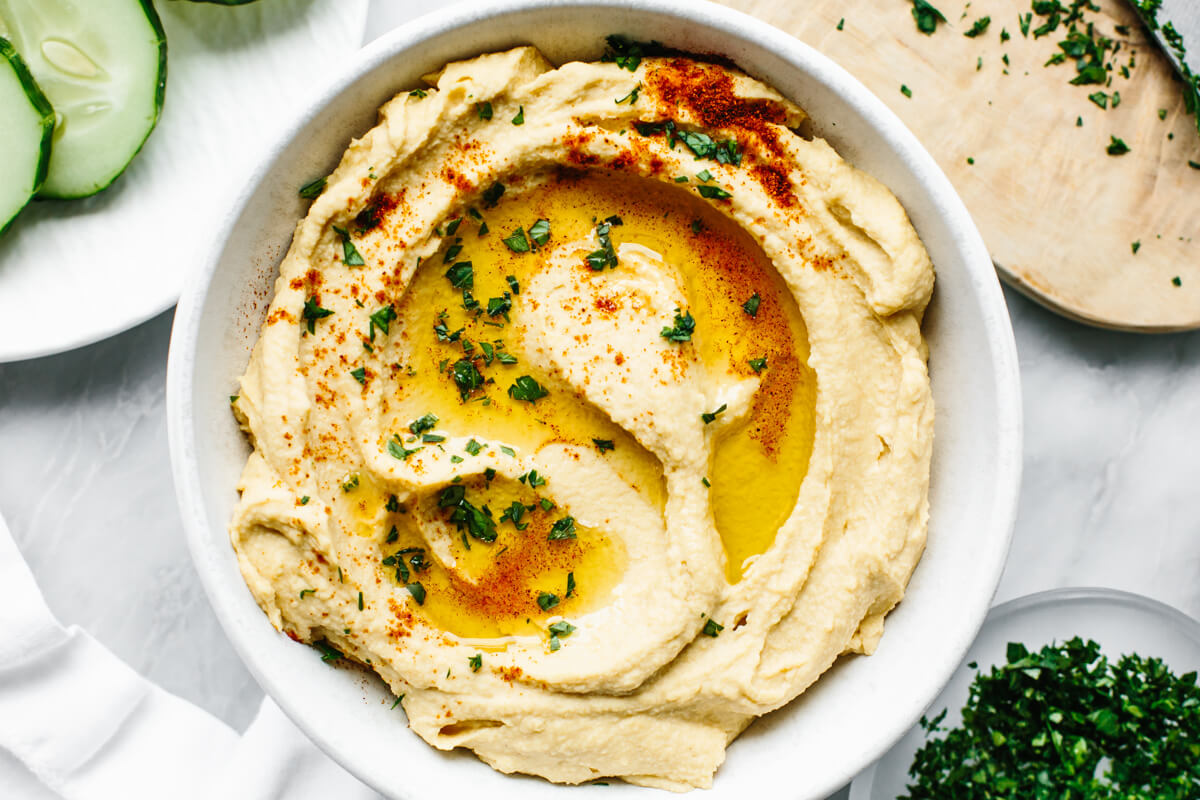 A white bowl of hummus next to chopped parsley