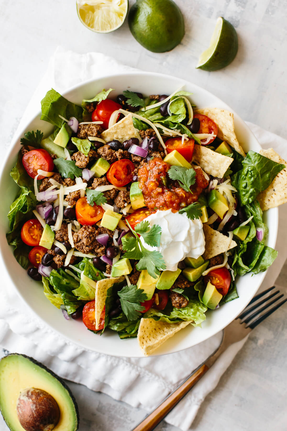 Taco salad in a white bowl on top of a napkin, with limes and half an avocado next to the bowl.