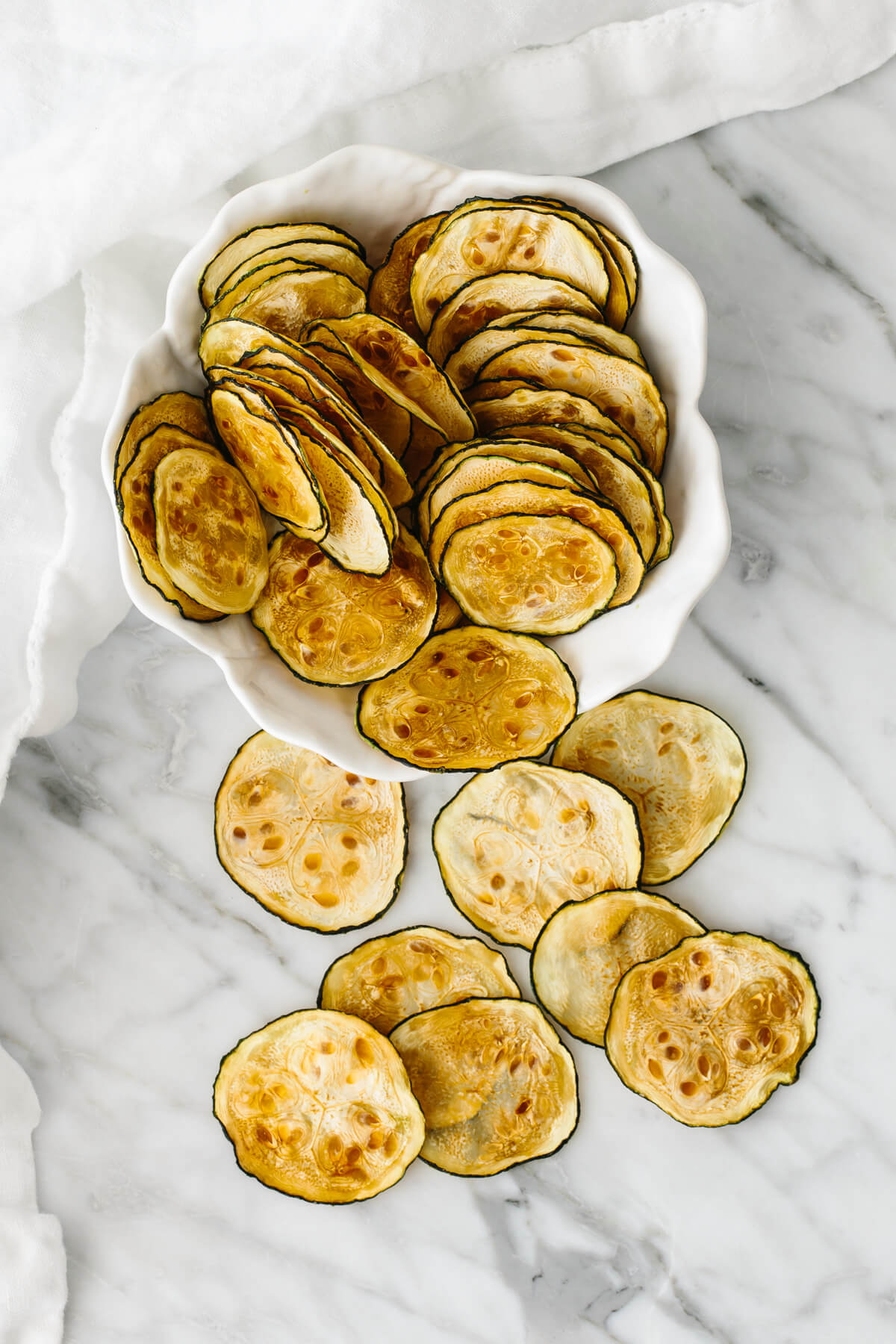 A white bowl with zucchini chips and a few extra chips laying on the table.