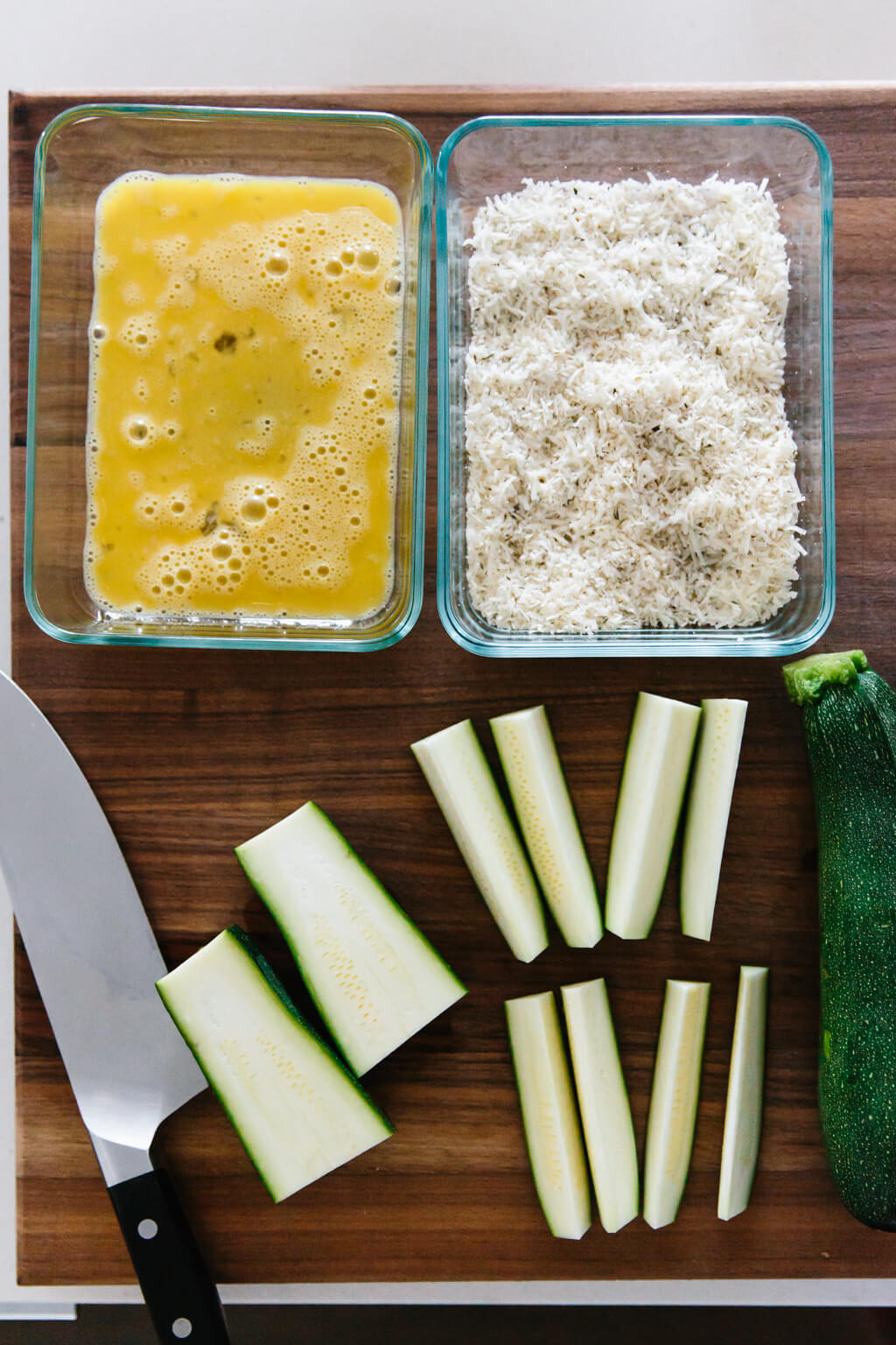 A bowl of egg wash and a bowl of parmesan cheese next to slices of zucchini. 